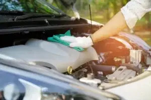 Motorwäsche: Person reinigt den Motorraum eines Autos mit Mikrofasertuch und Handschuhen bei geöffneter Motorhaube im Freien